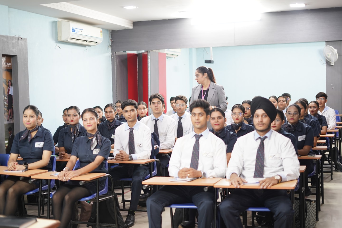 Students in uniforms sit at desks while an instructor observes.
